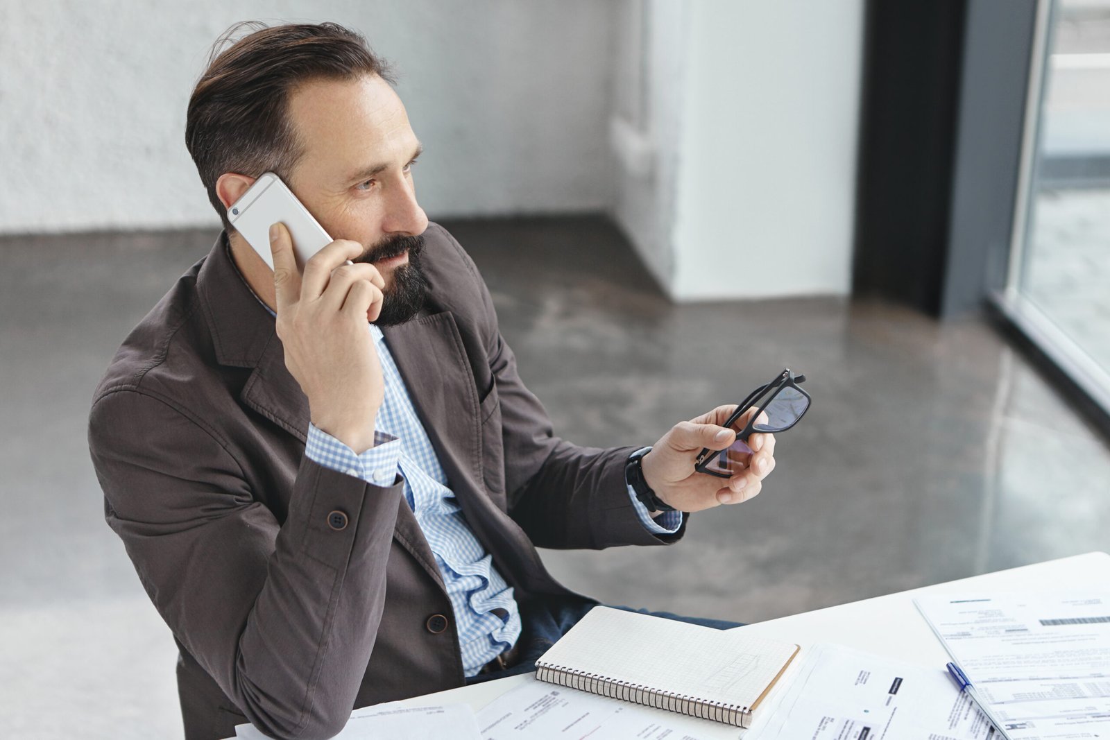Thoughtful businessman thinks about offer of collaboration, talks on mobile phone with mobile phone, has consultancy, works at documents, checks money on banking account, speaks with operator